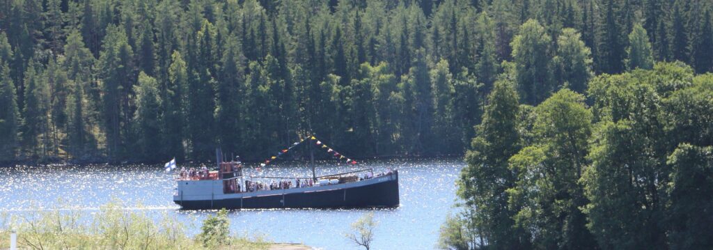 Vanha laiva seilaa kimmeltävällä järvelle ja taustaa vihreää metsää.