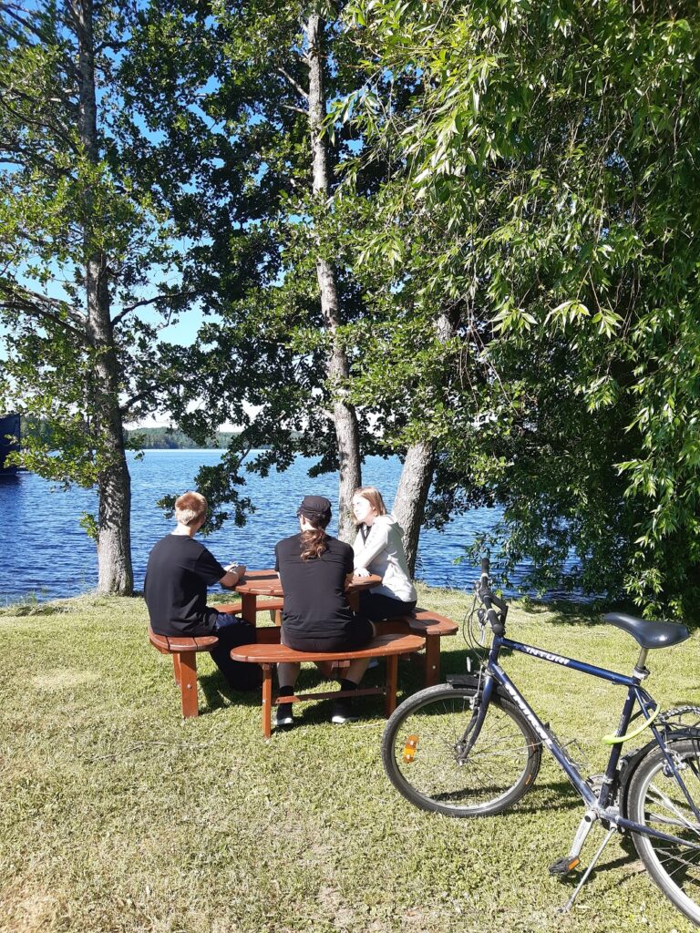 Kolme nuorta aikuista istuu ruskean pöytä-penkki-ryhmän ääressä. Vieressä polkupyörä, puita sekä järvenranta