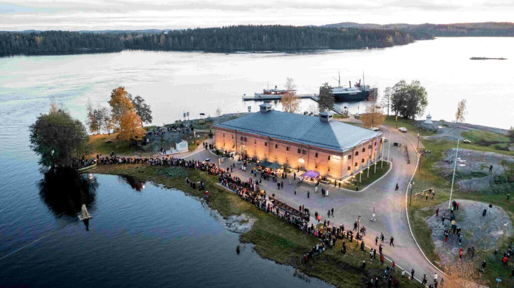 Keskellä museon vanha tiilinen rakennus ja ympärillä syksyinen järvimaisema vanhoine museolaivoineen. Rakennuksen etupihalla on suuret joukot ihmisiä sekä muutama myyntikoju.