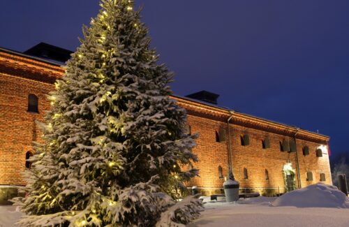 Joulukuusi valaistun tiilisen museorakennuksen edessä. Hämärä iltataivas taustalla sekä lumihankea.