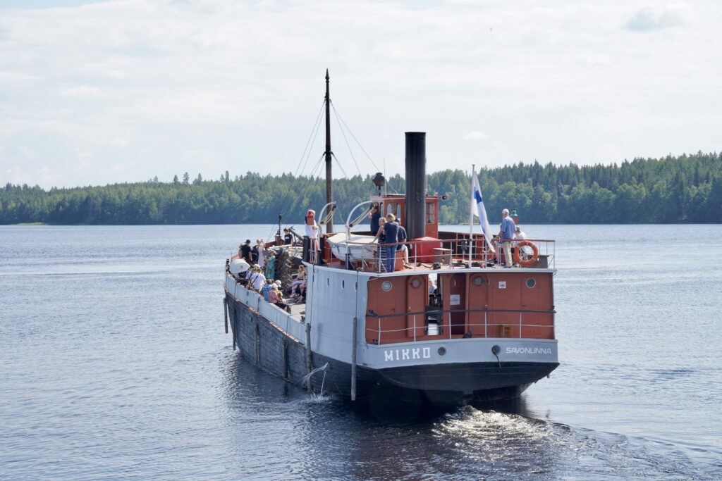 Vanha puna-harmaa-musta laiva kuvattu perästä päin. Seilaa kesäkelissä järvellä ja kyydissä on ihmisiä.