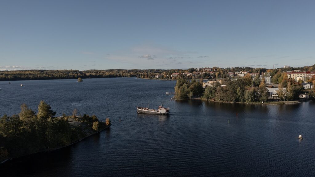 Vanha suuri laiva seilaa tummansinisellä järvenselällä. Oikealla näkyy kaupunkikeskustaa, vasemmalla laivan kulkusuunnassa pelkkää vettä, saaria ja metsää.